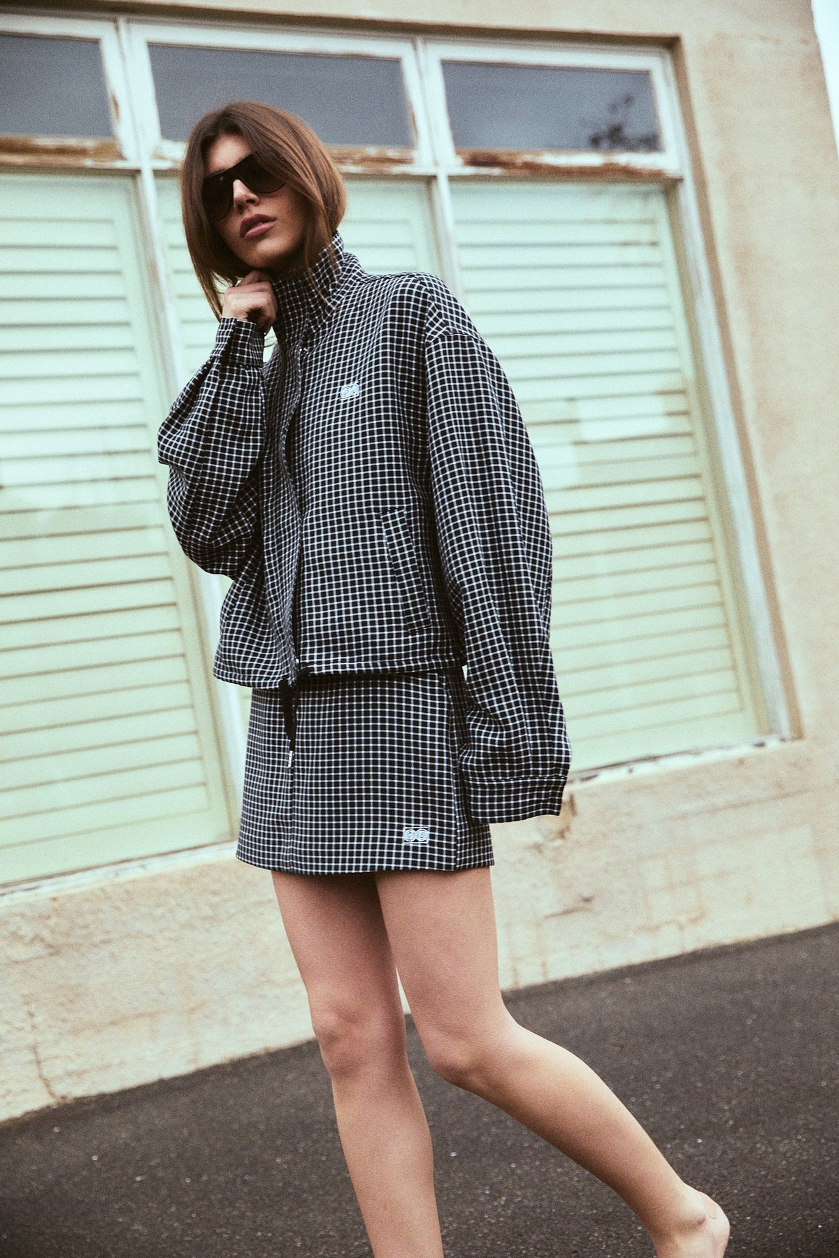 A model poses outdoors in a black and white check bomber jacket and matching mini skort, with one hand raised near the collar to highlight the long sleeves, relaxed fit and coordinated set.