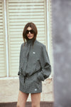 A model wearing a black and white check bomber jacket and matching mini skort poses outdoors in dark sunglasses, showing the coordinated pattern, high collar and relaxed shape of the jacket.