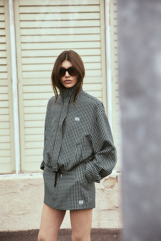 A model wearing a black and white check bomber jacket and matching mini skort poses outdoors in dark sunglasses, showing the coordinated pattern, high collar and relaxed shape of the jacket.