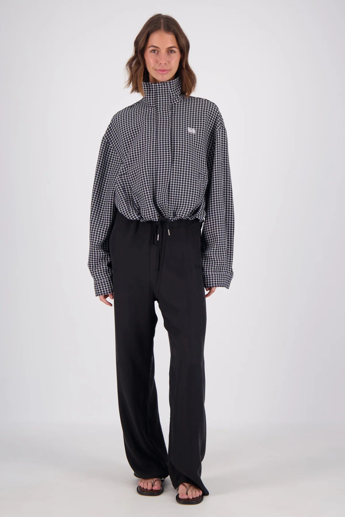  A model stands against a plain white background wearing a black and white check bomber jacket with a high neckline, long sleeves and gathered drawstring hem, paired with black wide-leg trousers and flat sandals.