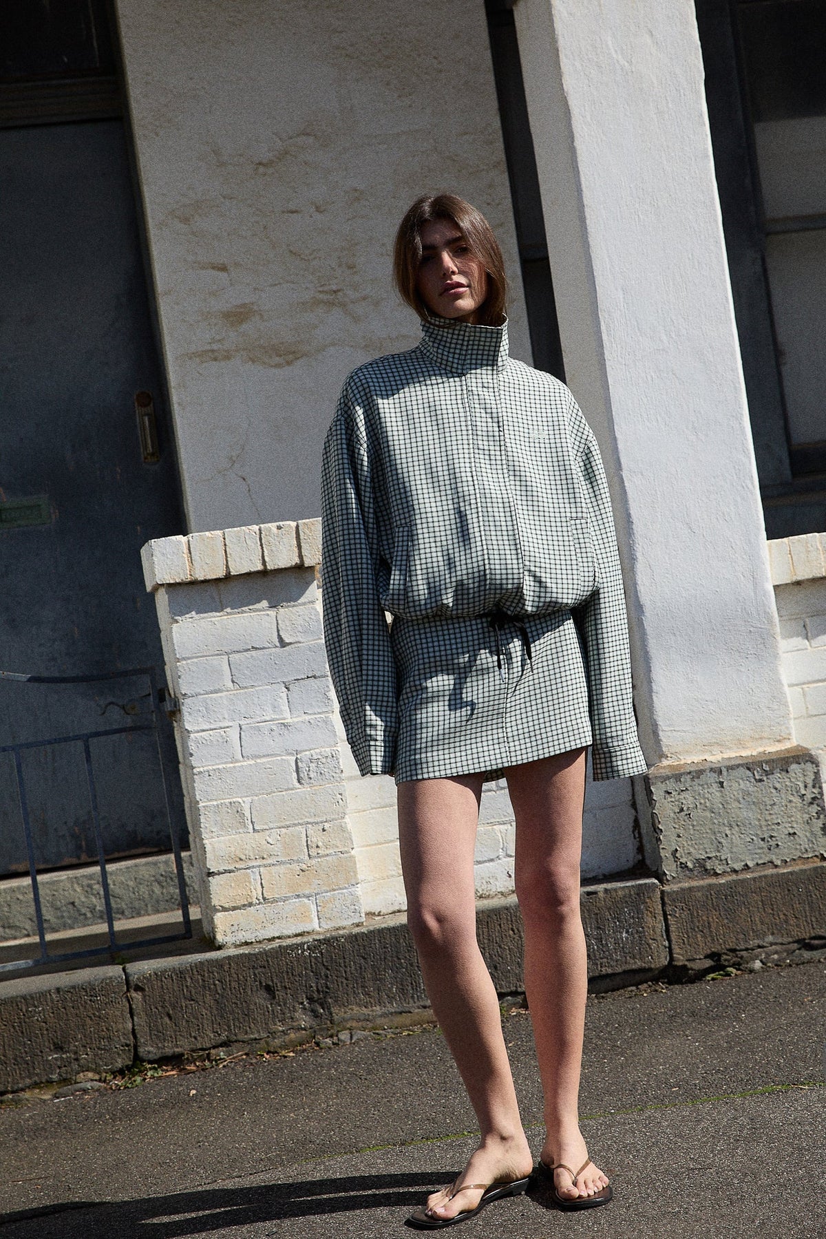 A full-length angled outdoor view of the white and mint check bomber jacket paired with a matching mini skirt, showing its relaxed shape, long sleeves and adjustable drawstring hem.