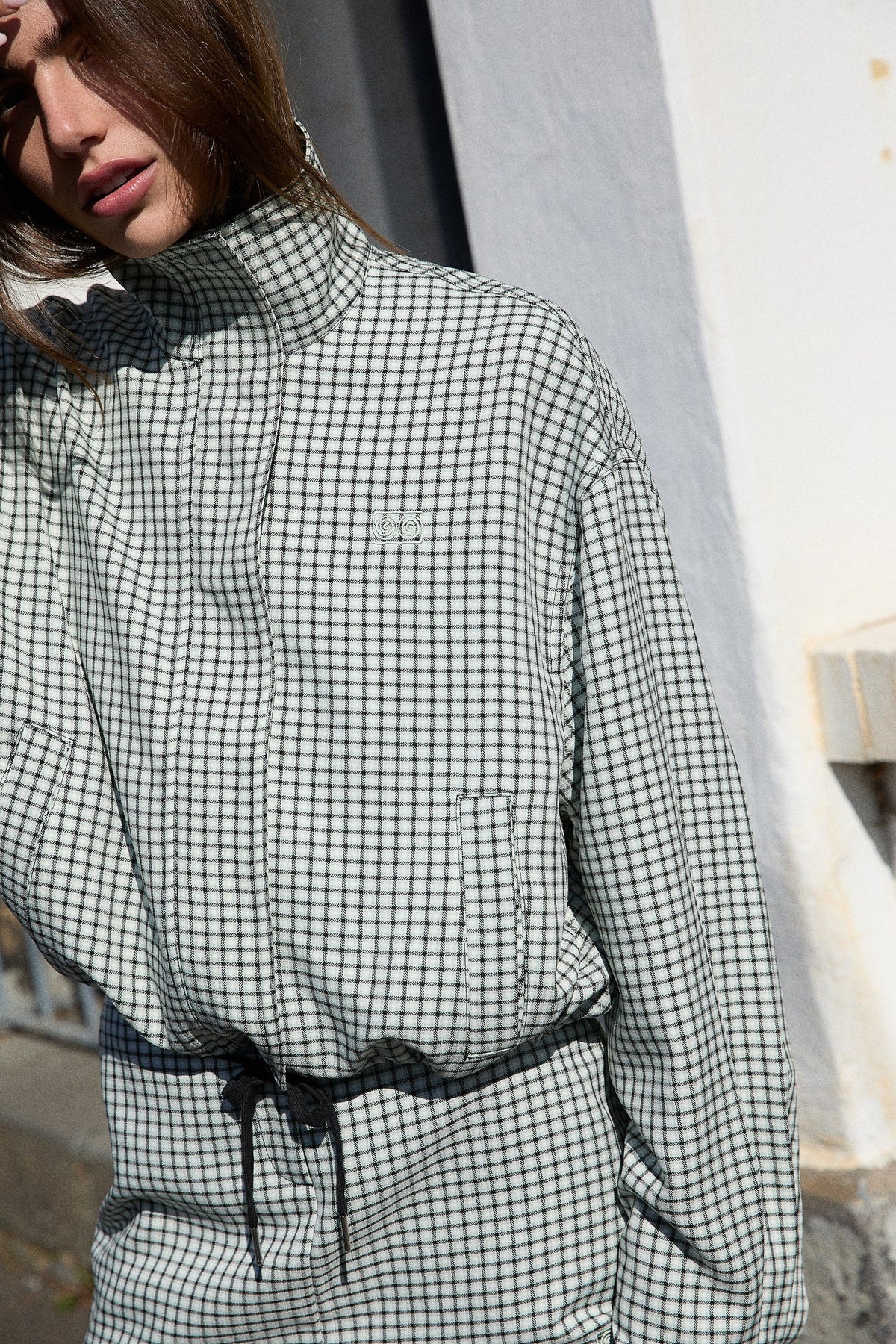 A close-up outdoor view of the white and mint check bomber jacket, highlighting the fine grid pattern, raised collar, front fastening and small embroidered chest emblem.