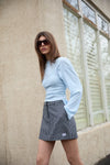 A model poses outdoors in the black and white check mini skort with a pale blue long-sleeved top and dark sunglasses, highlighting the check pattern, short hemline and small embroidered logo near the lower edge.