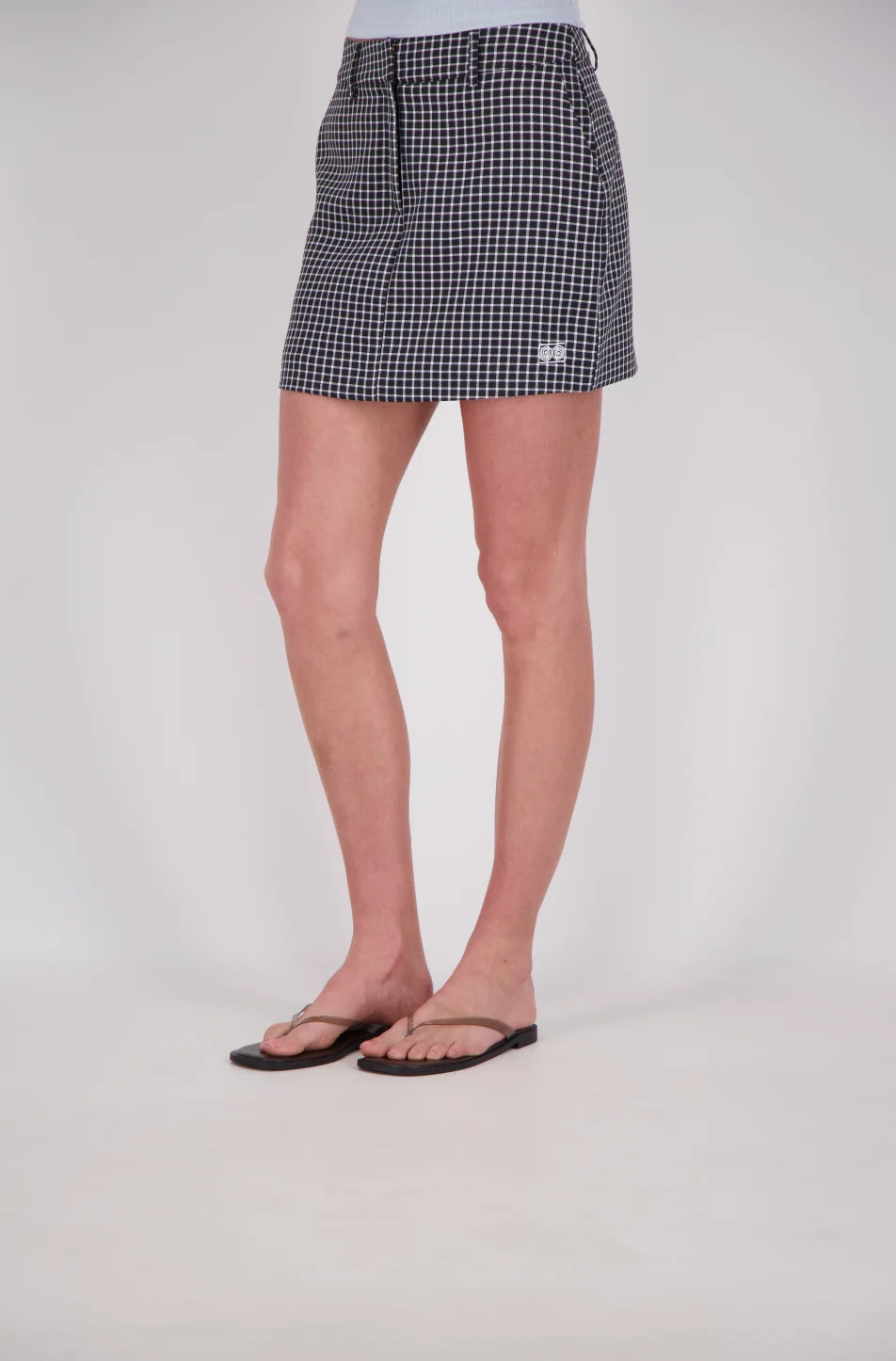 Front three-quarter view of a black and white check mini skort with a structured shape, belt loops, front pockets, a concealed front fastening and a small embroidered double-circle logo near the hem.