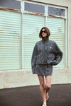 A model wears a black and white check mini skort with a matching high-neck jacket, long sleeves and flat sandals, standing outdoors in front of pale green window shutters.