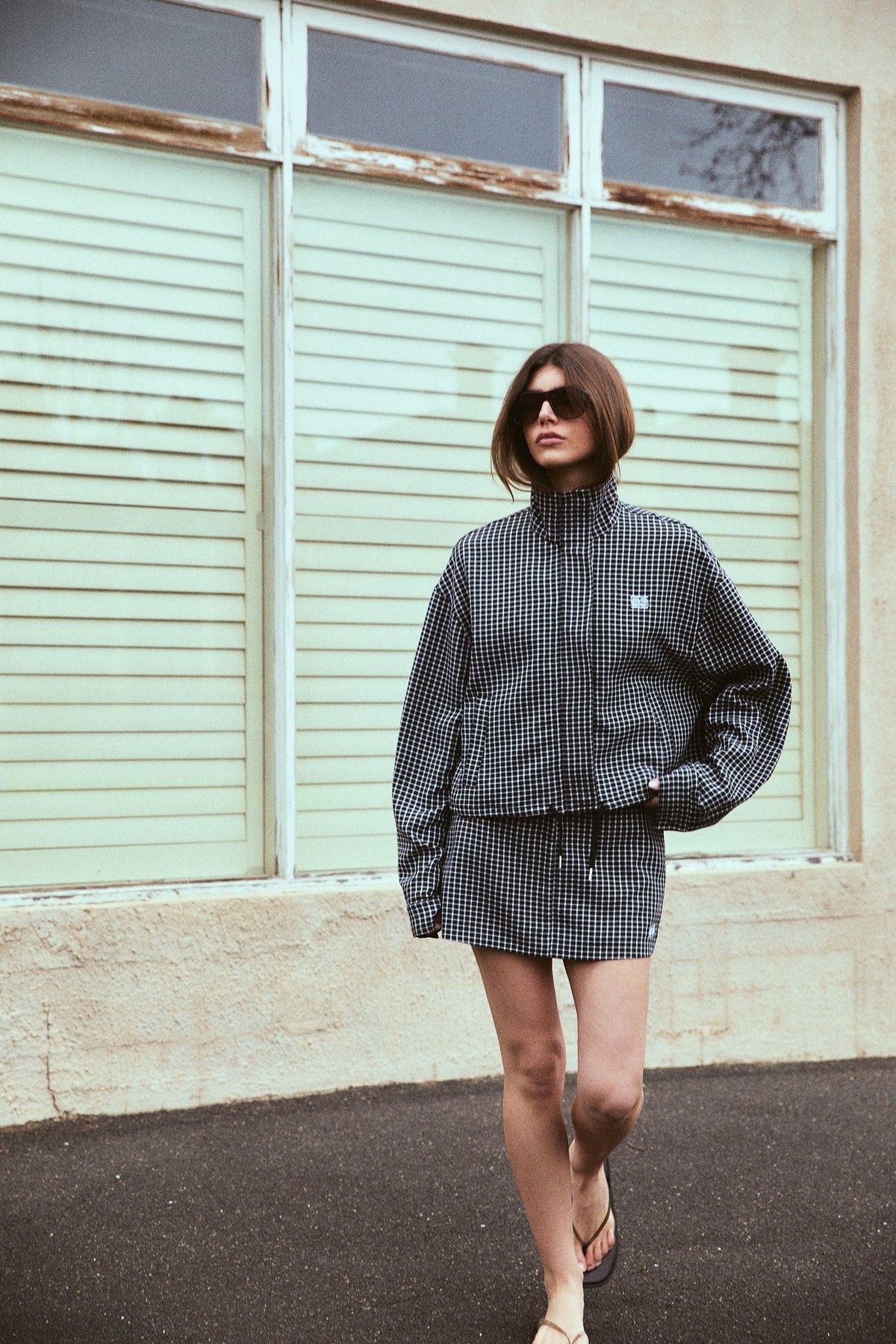 A model wears a black and white check mini skort with a matching high-neck jacket, long sleeves and flat sandals, standing outdoors in front of pale green window shutters.