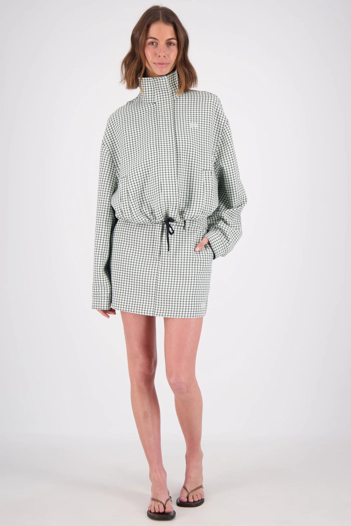 A woman modelling a white and mint checked outfit with a high-neck zip jacket and matching mini skort, photographed from the front in a studio.