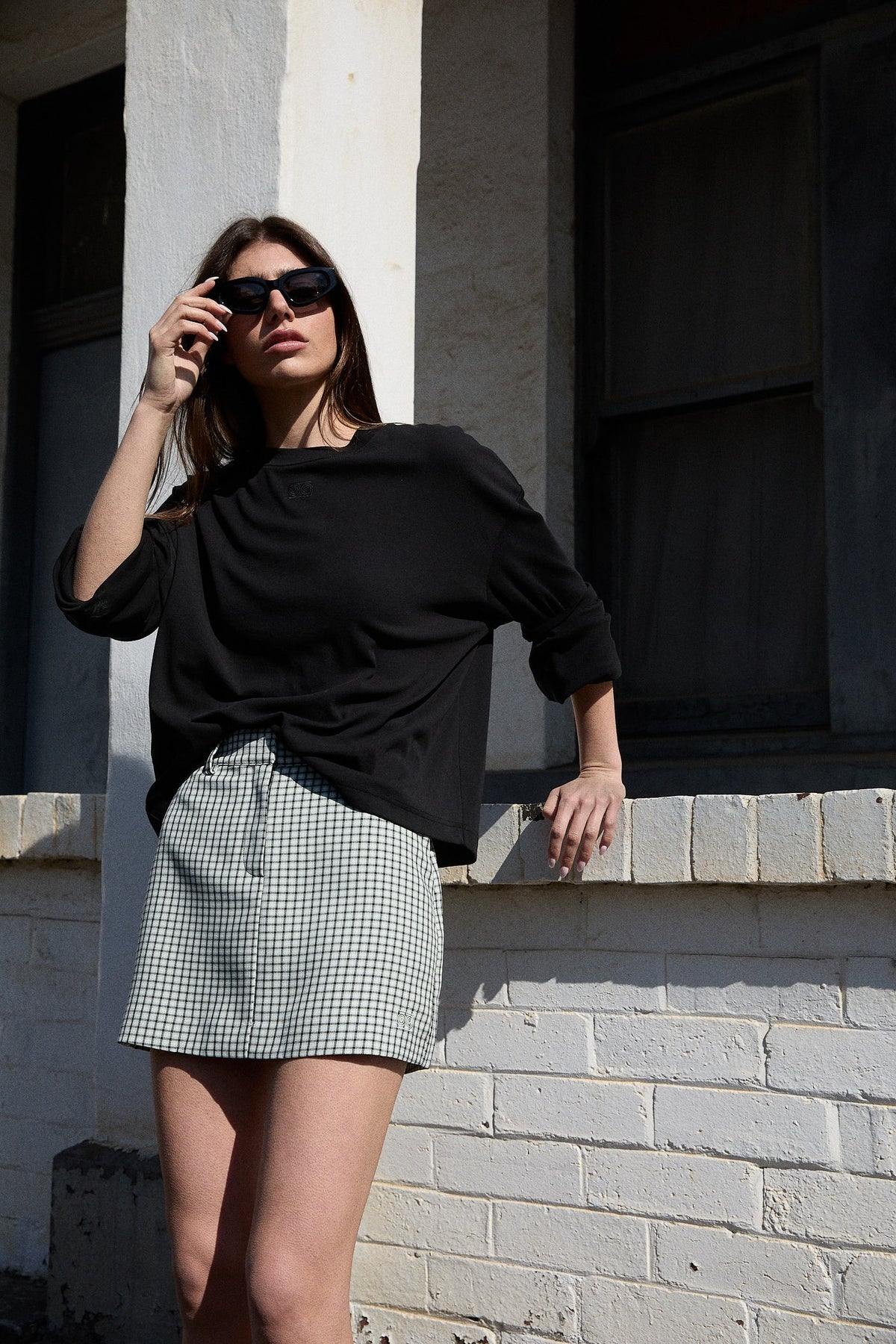 A woman wearing a white and mint checked mini skort with a black long-sleeve top and dark sunglasses, standing outdoors against a light brick wall.