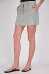 A front three-quarter view of a white and mint checked mini skort with a structured waistband, belt loops, front pockets and a small embroidered logo near the hem, styled with a black top and flat sandals.