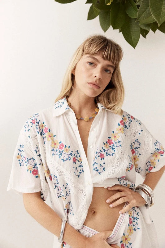 A woman standing in a bright studio, looking directly at the camera with one hand on her hip and the other resting near her skirt waistband. She models a white collared shirt with short sleeves, button fastening, colourful floral embroidery and lace detailing, paired with a matching white embroidered mini skirt.