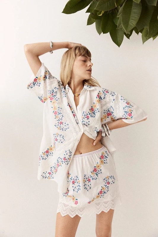 A woman posing in a bright studio with one arm raised above her head and the other resting on her hip. She wears a white short-sleeve button-up shirt featuring colourful floral embroidery, teal leaves, purple vines and decorative lace panels, styled with a matching mini skirt and silver bangles.