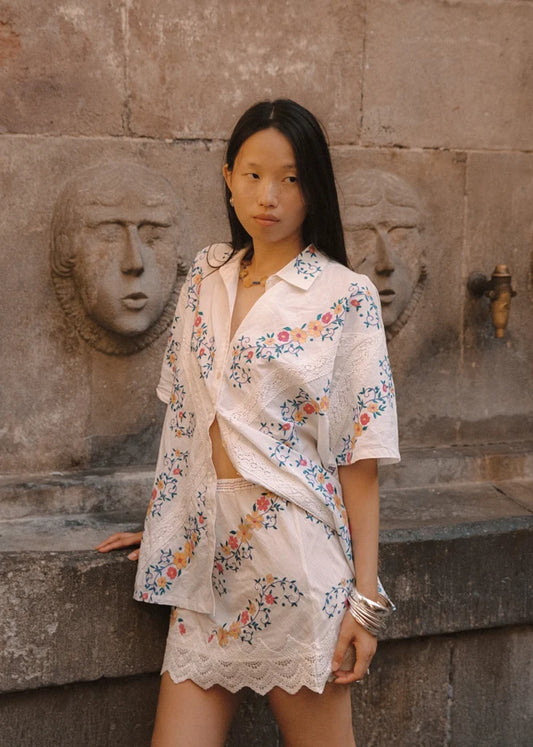 A woman posing in front of a carved stone wall, looking to the side with one hand resting on a ledge. She wears a white mini skirt with colourful floral embroidery and a scalloped lace hem, styled with a matching short-sleeve shirt and silver bangles.