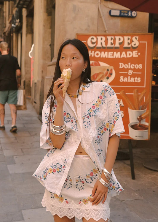 A woman standing on a European-style street while eating an ice cream cone and looking to the side. She wears a white mini skirt with colourful floral embroidery and a scalloped lace hem, paired with a matching short-sleeve shirt, silver bangles and dark sandals.
