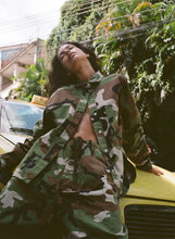 Person wearing a camouflage jacket leaning against a yellow car with greenery in the background