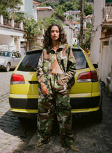 Person wearing a camouflage outfit standing in front of a yellow car on a street.