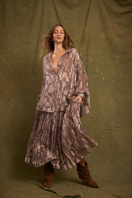 A model wears the purple metallic floral shirred maxi skirt with a matching oversized shirt and brown boots against an olive studio backdrop.