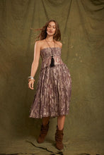 A model wears the purple metallic floral shirred maxi skirt with a coordinating strapless top and brown boots against an olive studio backdrop.