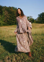 A model wears the flowing purple metallic floral maxi skirt with a matching shirt while posing outdoors in a sunlit grassy field.