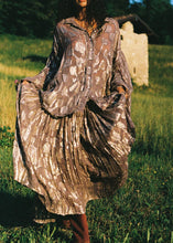 A model holds the sides of the purple metallic floral maxi skirt to show its gathered volume while standing outdoors in a grassy field.