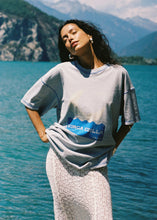 Model wearing the oversized grey marl tee with a cream lace skirt by the water, showing the relaxed short sleeves and bold blue front graphic.