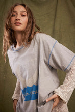 Close-up of the grey marl graphic tee layered over cream lace sleeves, showing the blue contrast stitching, relaxed neckline and faded front artwork.