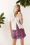 A woman standing in a bright studio, looking toward the camera while holding the collar of her white short-sleeve shirt. She pairs the shirt with a plum mini skirt featuring dark polka dots and a wide scalloped lace hem, along with silver jewellery.