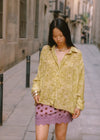 A woman walking along a stone-paved street with her head lowered, holding the hem of her oversized yellow-green patterned shirt. She wears a plum mini skirt with dark polka dots and a scalloped lace hem, accessorised with silver bangles.