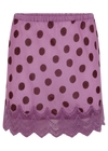 A close-up of a plum mini skirt displayed against a black background, featuring dark polka dots, an elasticated waistband and a wide lace trim with intricate patterns and a scalloped edge.
