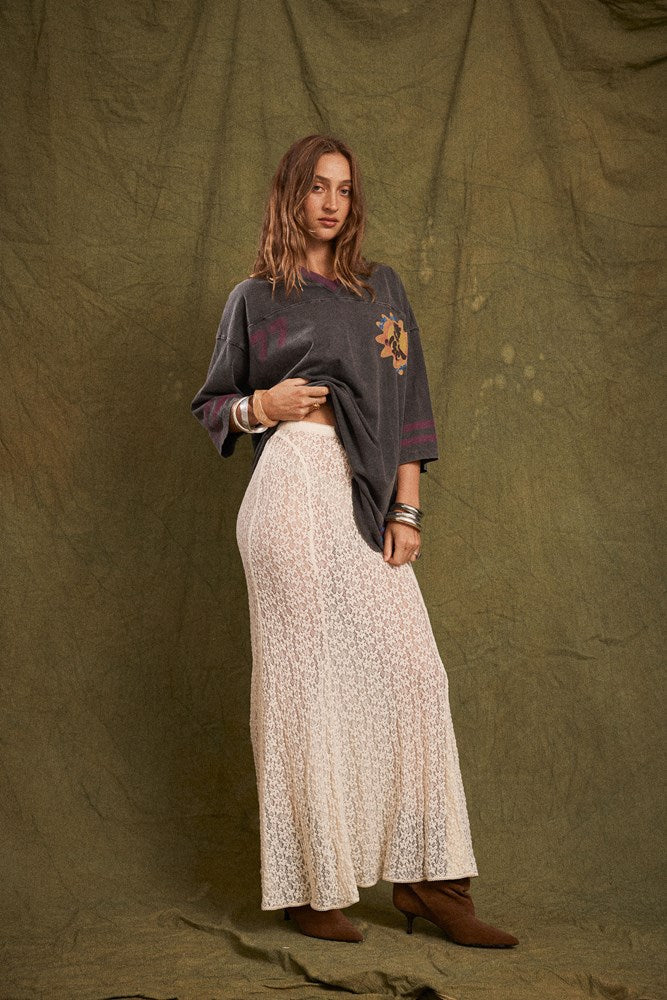 Model wearing the cream lace maxi skirt with an oversized graphic tee and brown ankle boots, showing the long flowing shape, delicate lace texture and gently flared hem in an angled front view.
