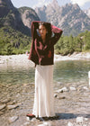 Model wearing the cream lace maxi skirt with a brown patterned cardigan and ankle boots beside a mountain lake, showing the full-length silhouette and intricate lace texture in a lifestyle front view.