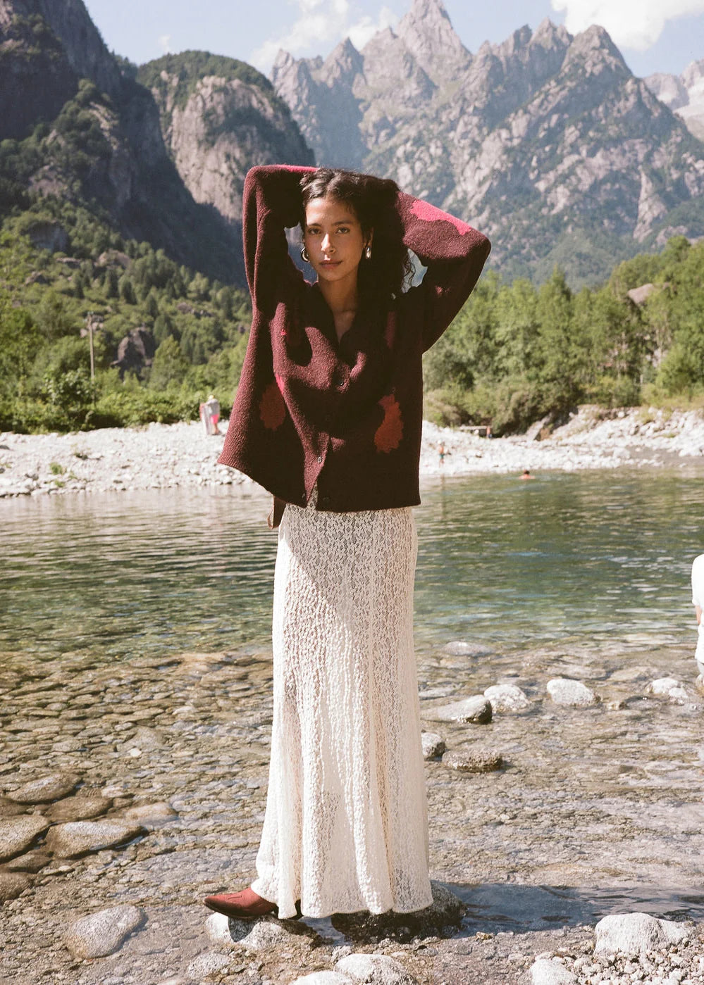 Model wearing the cream lace maxi skirt with a brown patterned cardigan and ankle boots beside a mountain lake, showing the full-length silhouette and intricate lace texture in a lifestyle front view.