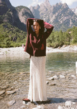 Model wearing the cream lace maxi skirt with a brown patterned cardigan and ankle boots beside a mountain lake, showing the full-length silhouette and intricate lace texture in a lifestyle front view.
