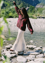 Model wearing the cream lace maxi skirt with a brown cardigan beside a mountain lake, showing the flowing maxi length, softly flared hem and delicate lace finish in a lifestyle front view.
