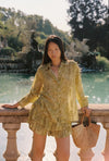 Lifestyle front view of the Dorato printed button-up shirt styled with matching ruffle shorts and a woven tote bag beside a garden pond.