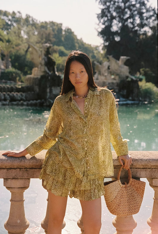 Lifestyle front view of the Dorato printed button-up shirt styled with matching ruffle shorts and a woven tote bag beside a garden pond.