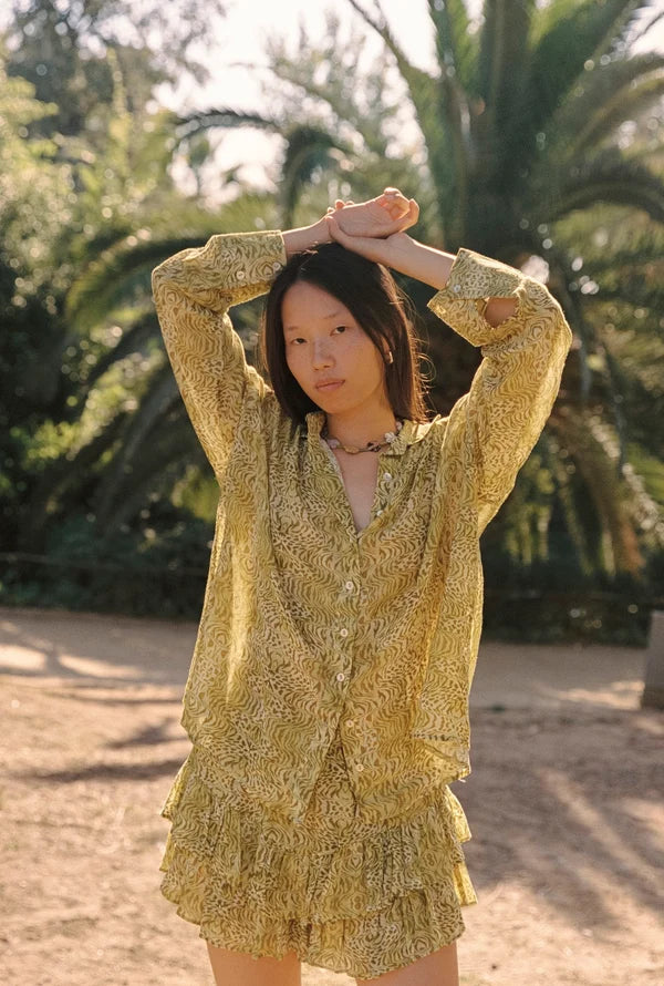 Lifestyle front view of the Dorato printed button-up shirt styled with matching ruffle shorts in a sunlit garden setting with palm trees.