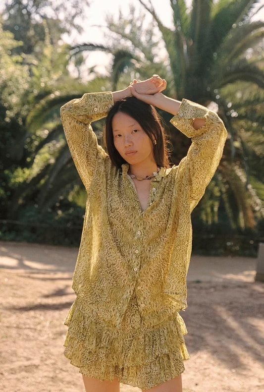Lifestyle front view of the Dorato printed button-up shirt styled with matching ruffle shorts in a sunlit garden setting with palm trees.