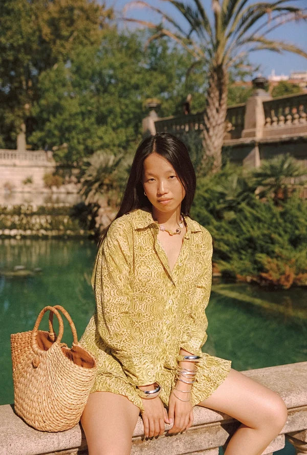 Lifestyle view of the Dorato printed button-up shirt styled with matching ruffle shorts while seated beside a garden pond with a woven tote bag.