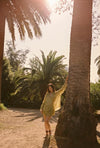 Lifestyle full-length view of the Dorato printed button-up shirt styled with matching ruffle shorts and black sandals beside a palm tree in a sunlit garden setting.