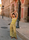 Lifestyle full-length front view of the Dorato printed wide-leg pants with a tasseled waist tie, styled with a light green tank top and black sandals on a sunlit street.