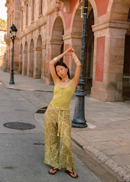 Lifestyle full-length front view of the Dorato printed wide-leg pants with a tasseled waist tie, styled with a light green tank top and black sandals on a sunlit street.