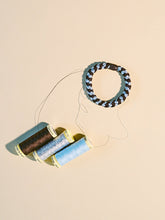 Brown and sea blue woven hair tie displayed beside matching thread spools on a neutral background.