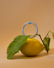 Pale silver-blue woven hair tie styled upright on a lemon with green leaves against a warm neutral background.