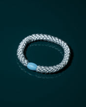 Baby Frost woven hair tie displayed flat on a dark teal surface with its light blue bead facing forward.