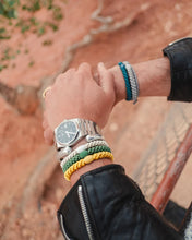 Forrest Green woven hair tie worn as a bracelet alongside stacked hair ties and a silver watch in an outdoor setting.