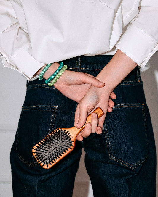 Lifestyle image of several green and teal woven hair ties worn around the wrist while holding a wooden hairbrush, styled with dark denim and a white top.