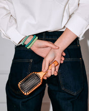 Lifestyle image of several green and teal woven hair ties worn around the wrist while holding a wooden hairbrush, styled with dark denim and a white top.