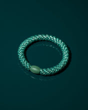 Close-up product shot of the teal glitter hair tie resting on a deep green surface, highlighting its woven texture, metallic sparkle and rounded bead.