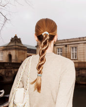 Slim teal and gold glitter hair tie securing a woman's braided ponytail outdoors in front of a historic building.