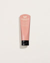 A pink 100ml hand balm tube with white lettering and a black flip-top cap is shown upright against a pale background with a soft shadow.